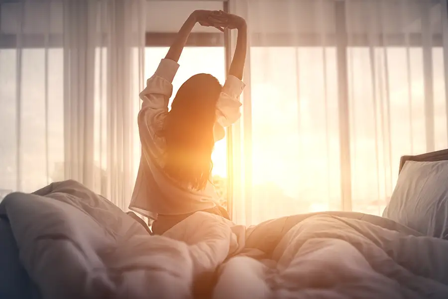 Woman sitting up in bed stretching her arms overhead as morning sunlight streams through sheer white curtains.